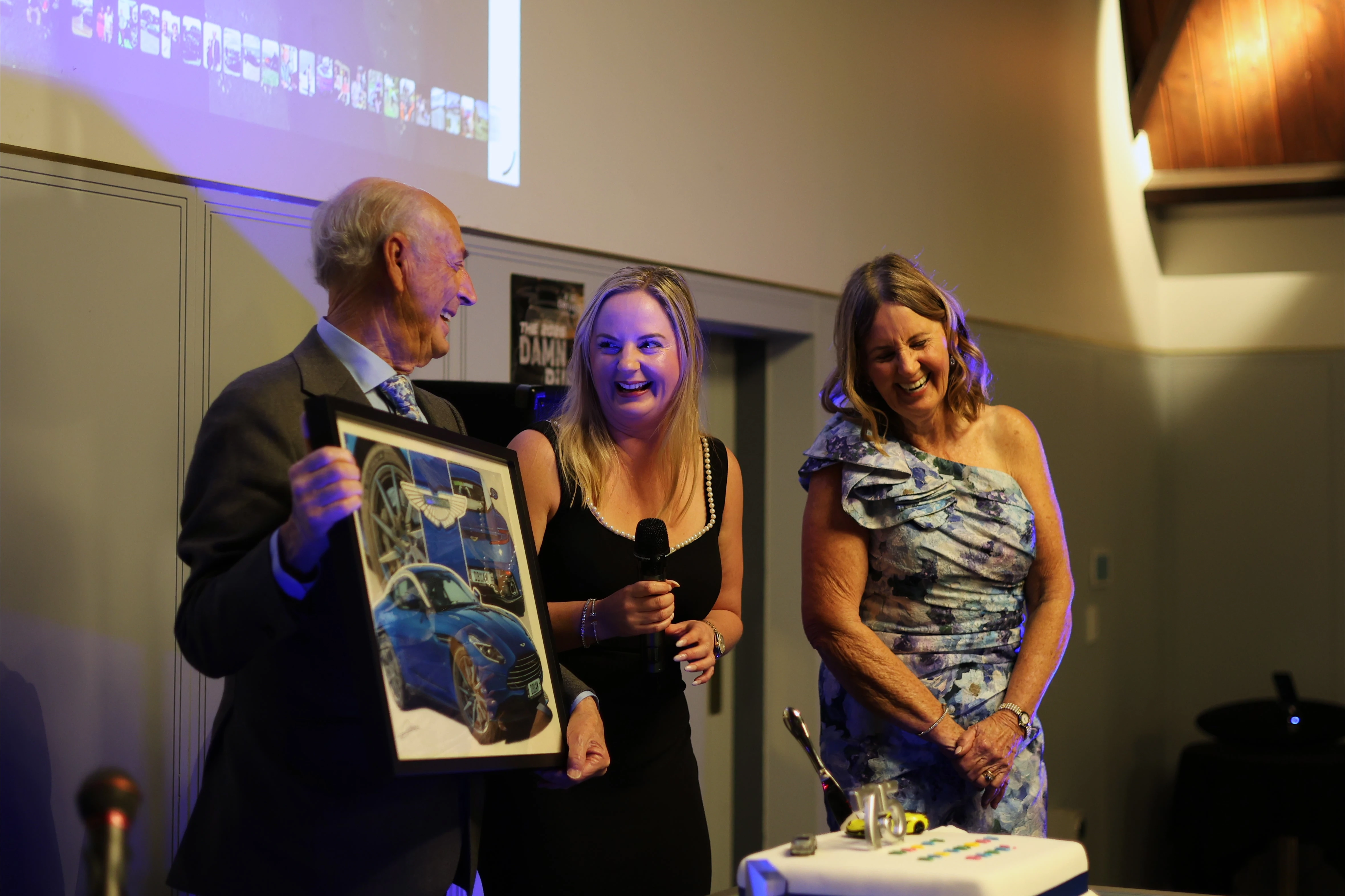 The birthday guest laughs with two women while holding a framed picture of cars