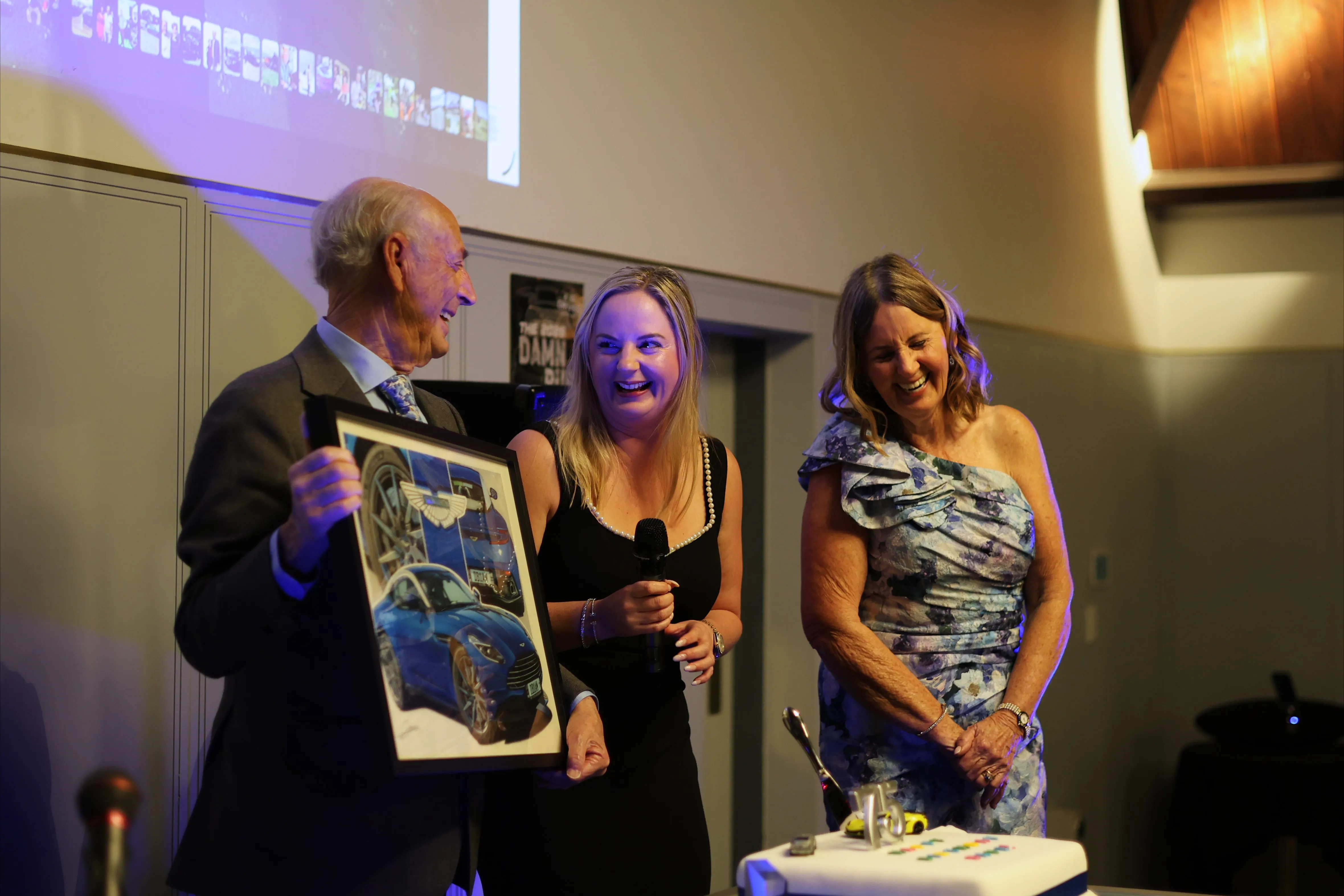 The birthday guest laughs with two women while holding a framed picture of cars