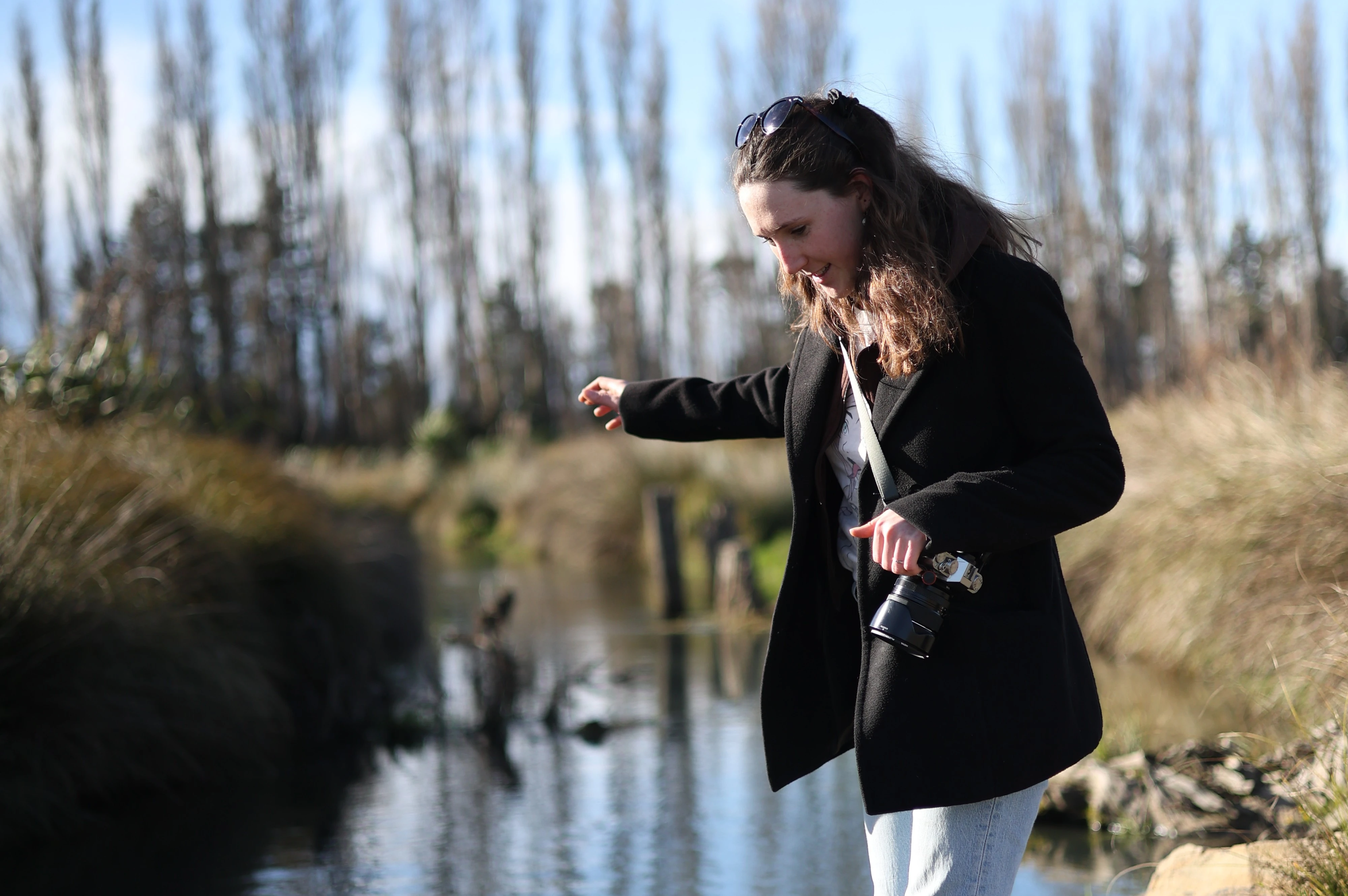 Caitlin walks beside a small stream with a camera in her hand on a bright sunny day
