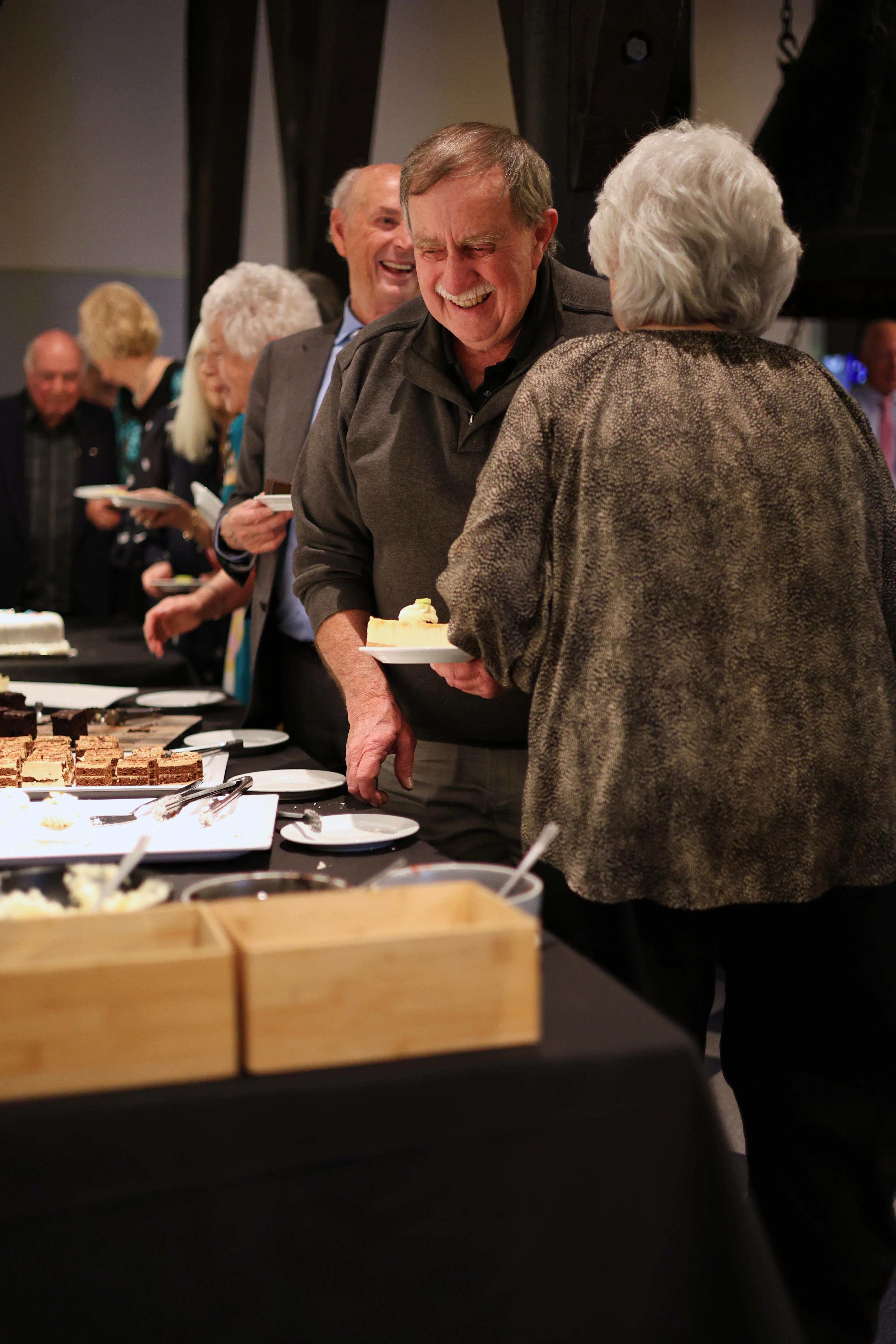 Two older guests smile at one another beside a table of desserts
