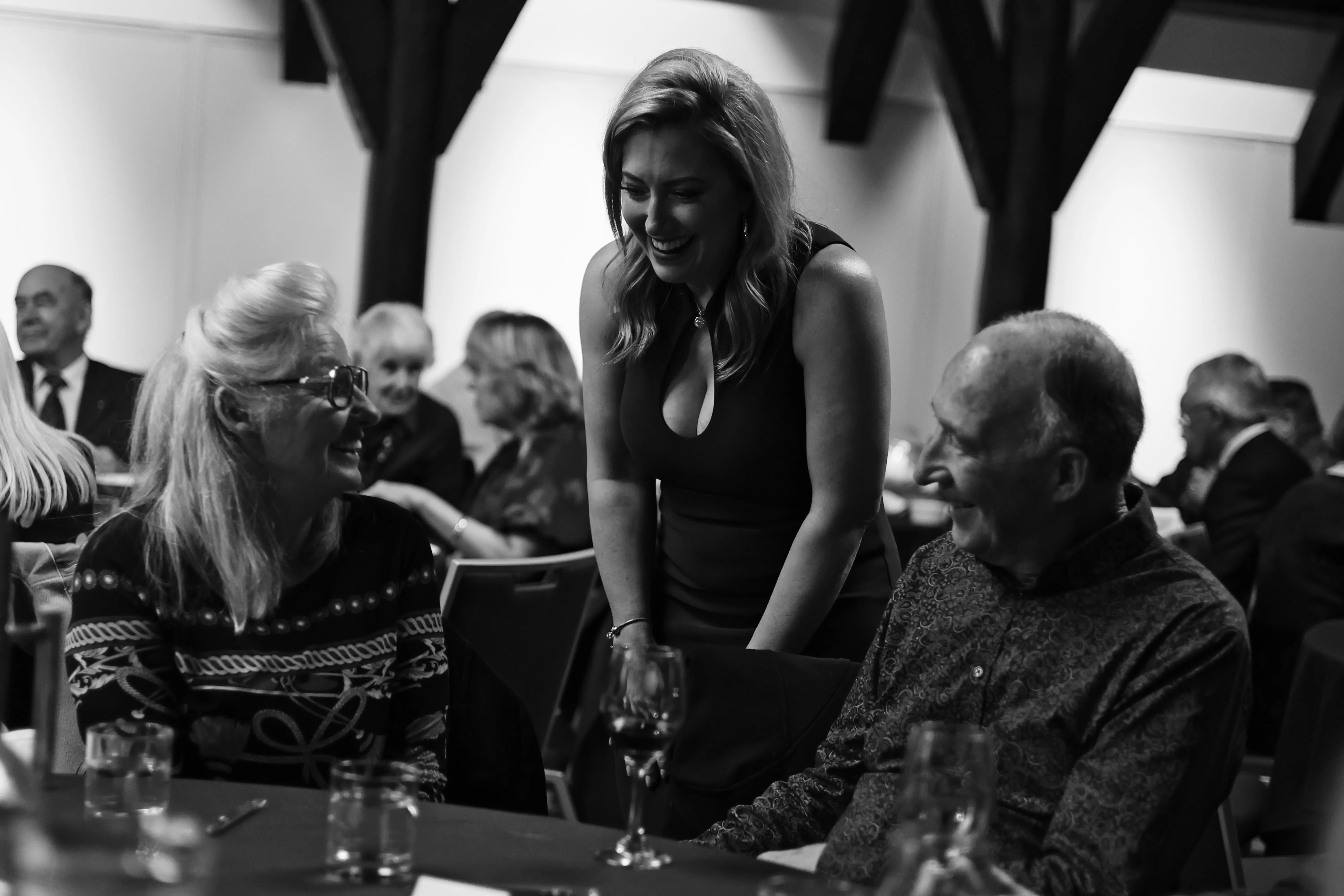 A woman in a black dress leans between seated guests and smiles