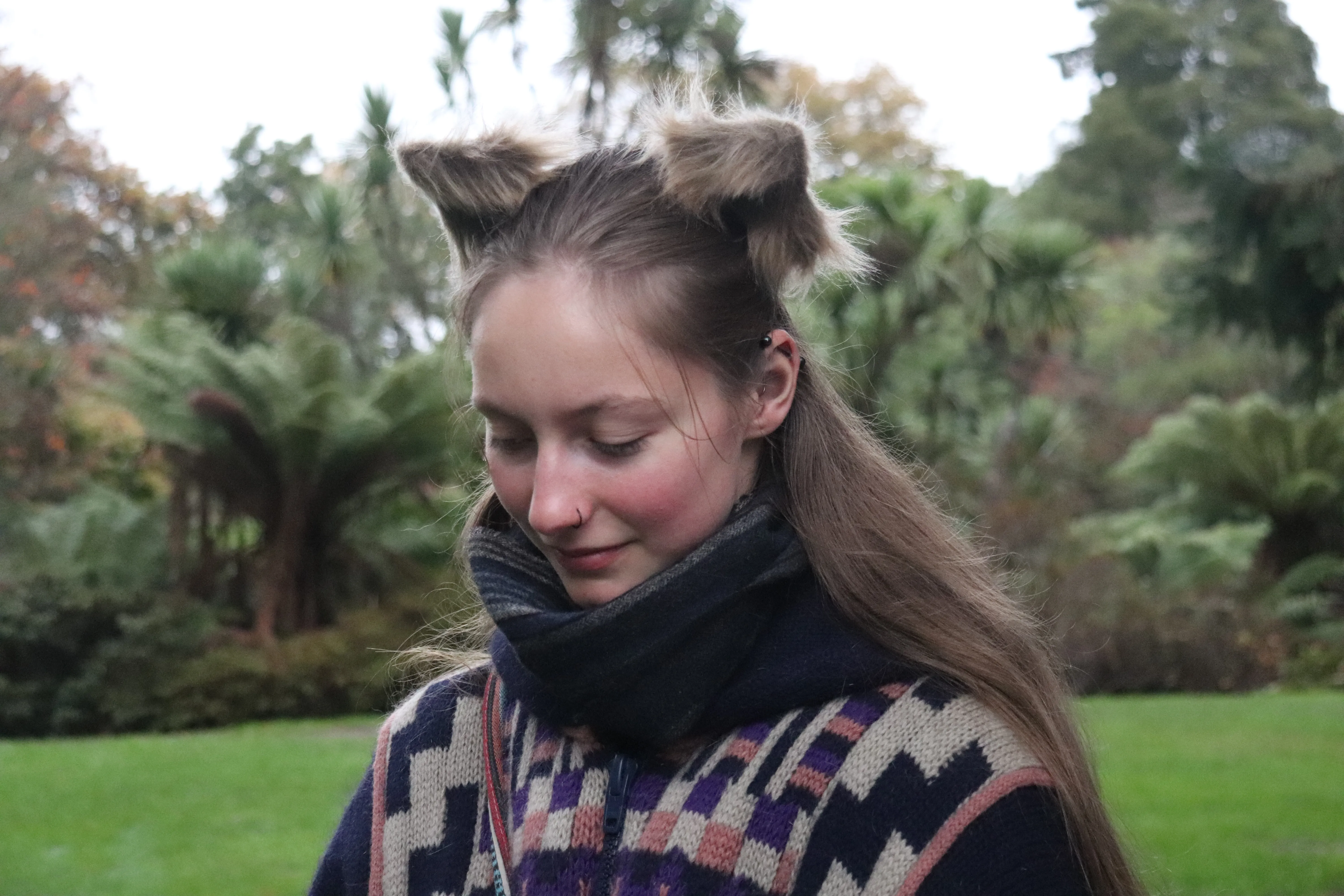 A woman with her hair styled into two pointed tufts looks down in a green garden