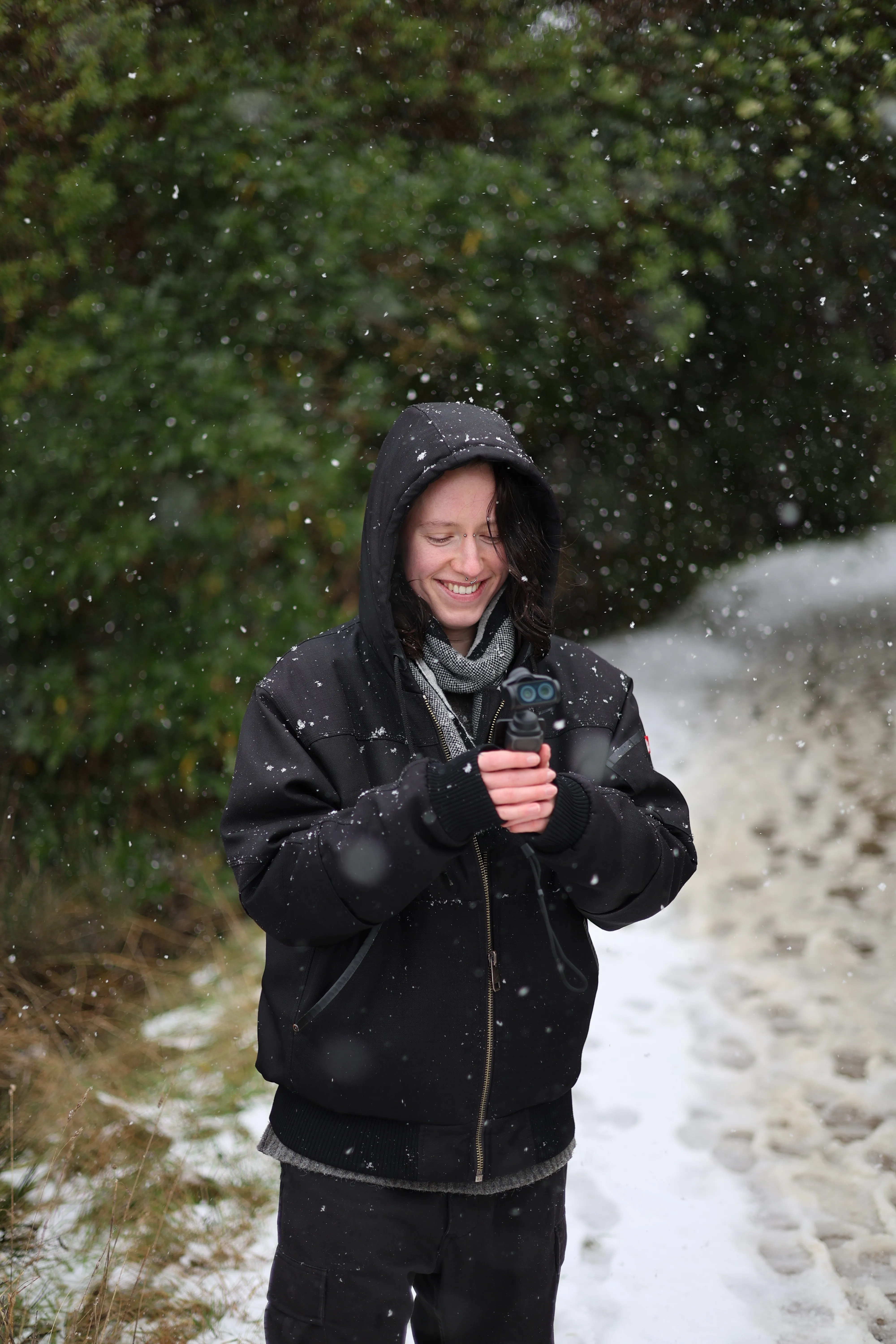 Haven smiles while holding a small camera as snow falls through the forest