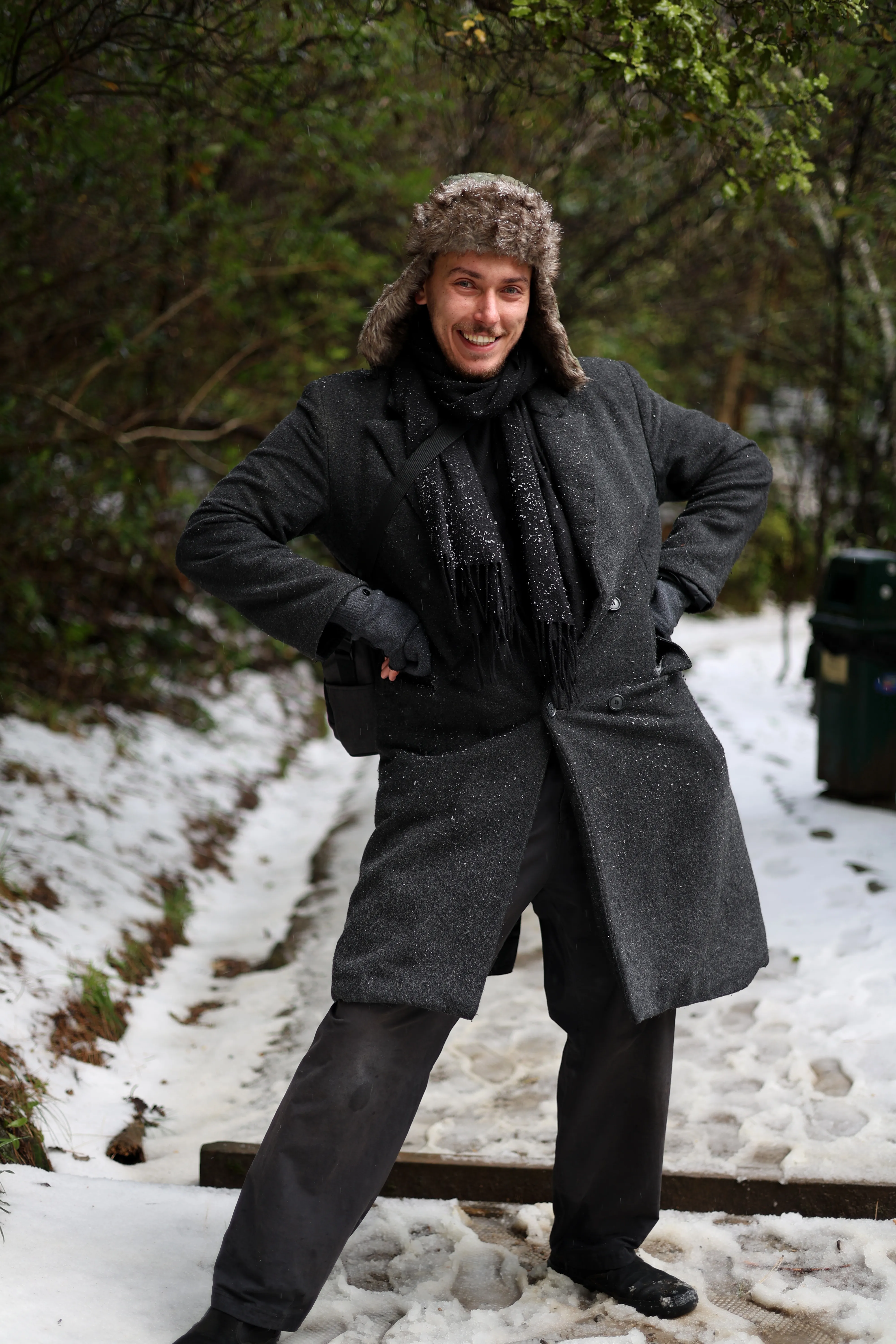 Kenan smiling with his hands on his hips, wearing a dark coat and fur hat on a snowy path