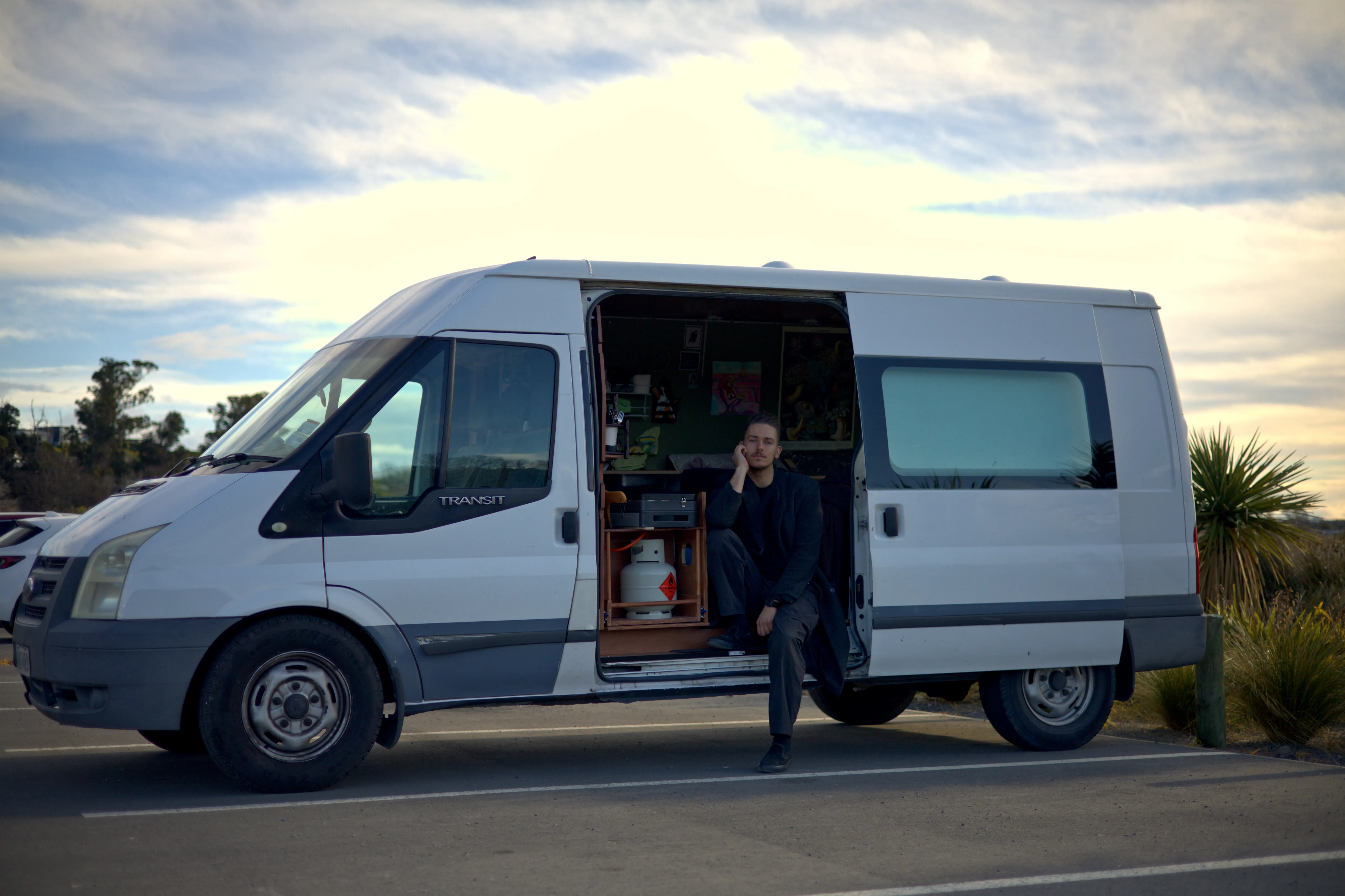 Kenan sitting on the open side step of his white van beneath a cloudy sky