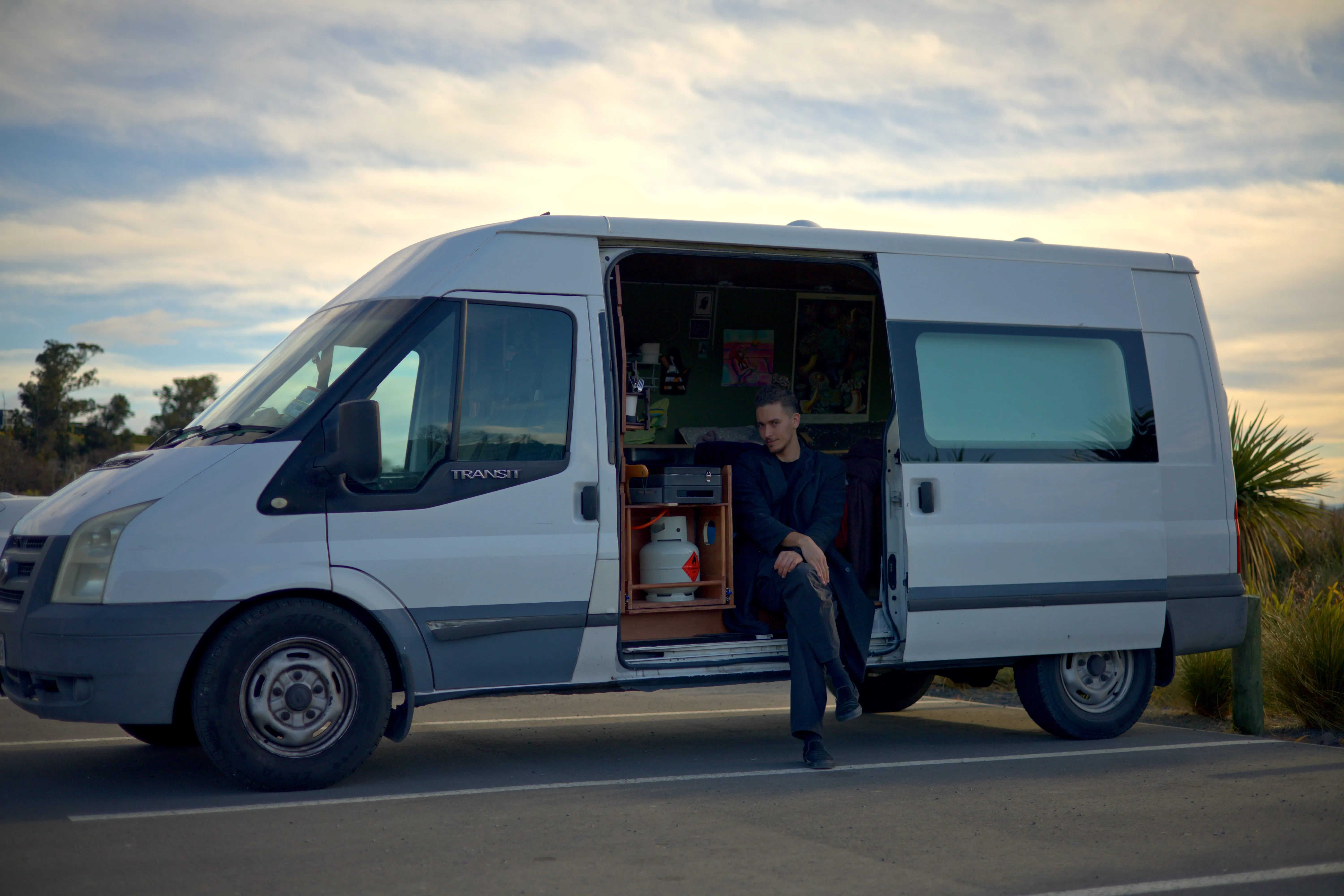 Kenan-John sitting on the open side step of his white van beneath a cloudy sky