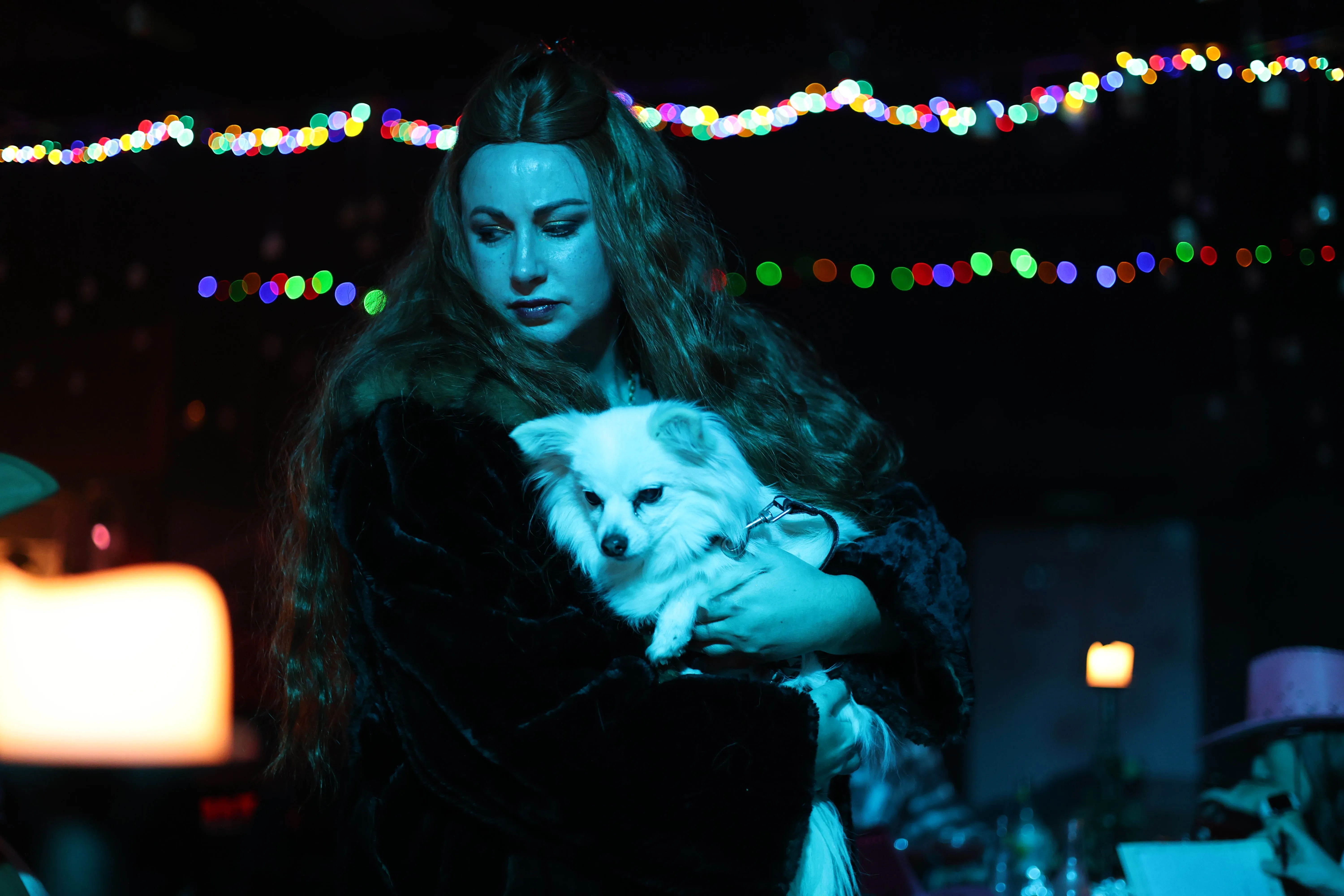 A woman in a dark costume holds a small white dog beneath strings of coloured lights