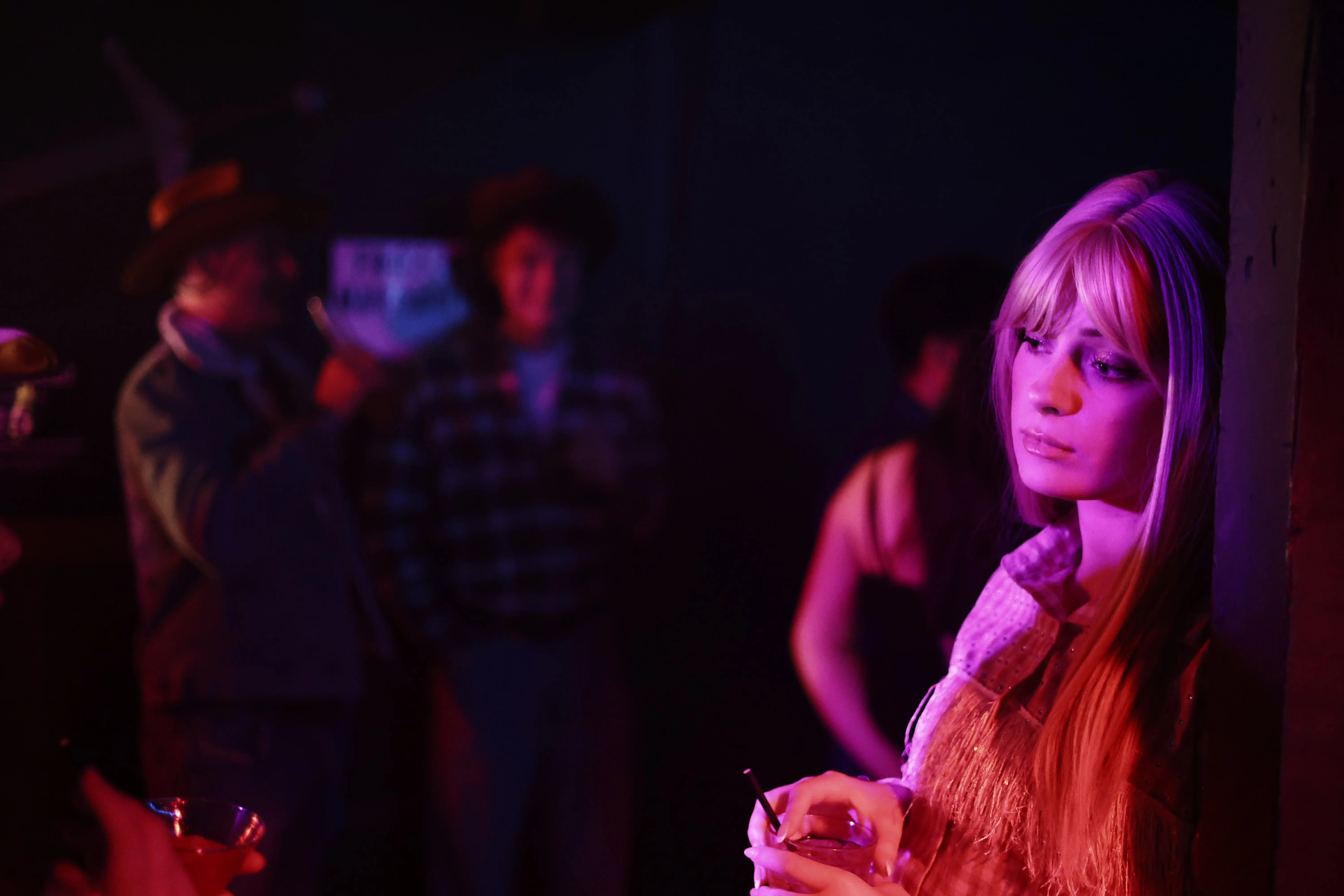 A woman in a blonde wig leans against a post under pink stage lighting