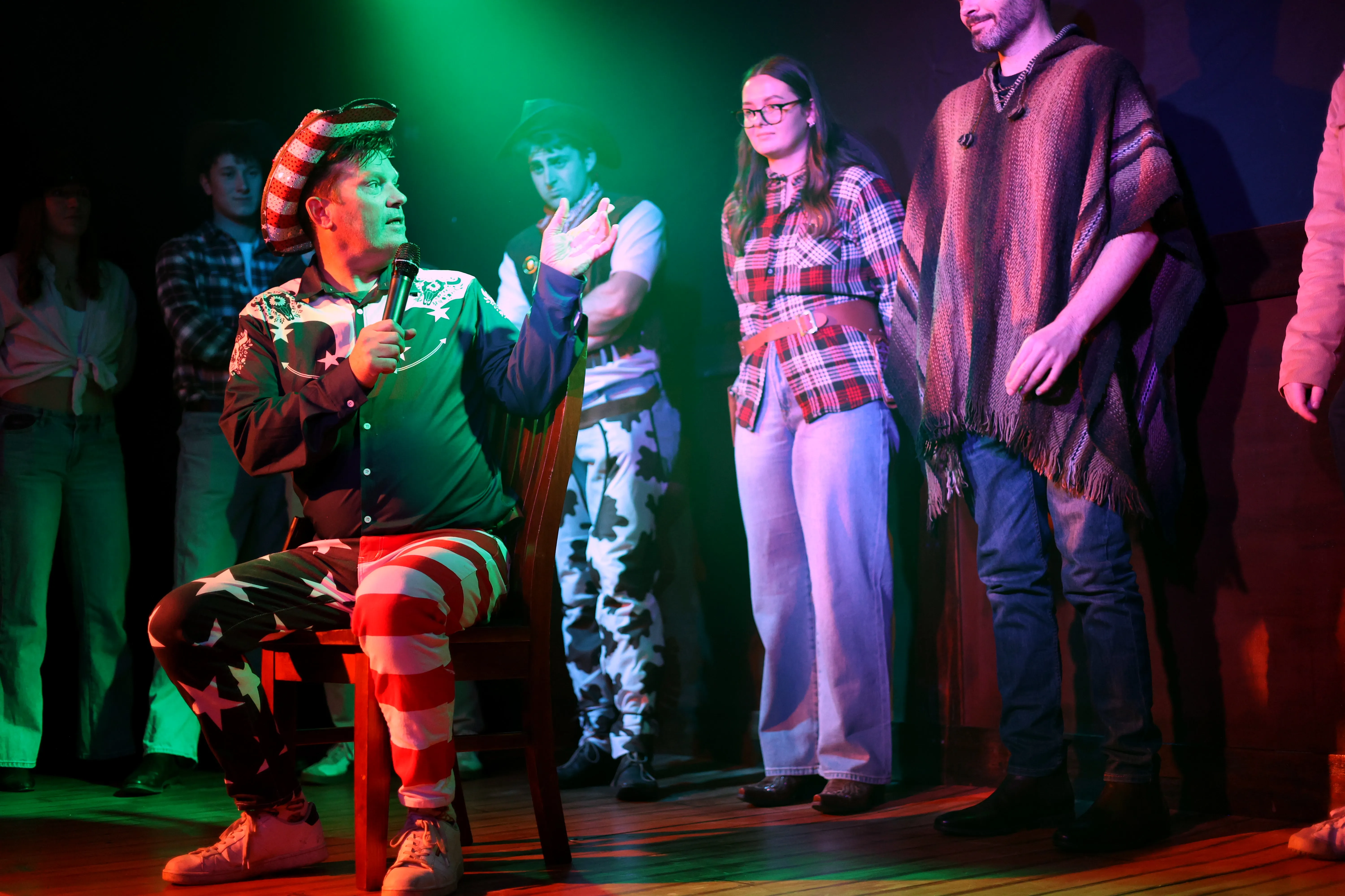 A man in a striped cowboy hat speaks into a microphone while costumed guests stand behind him