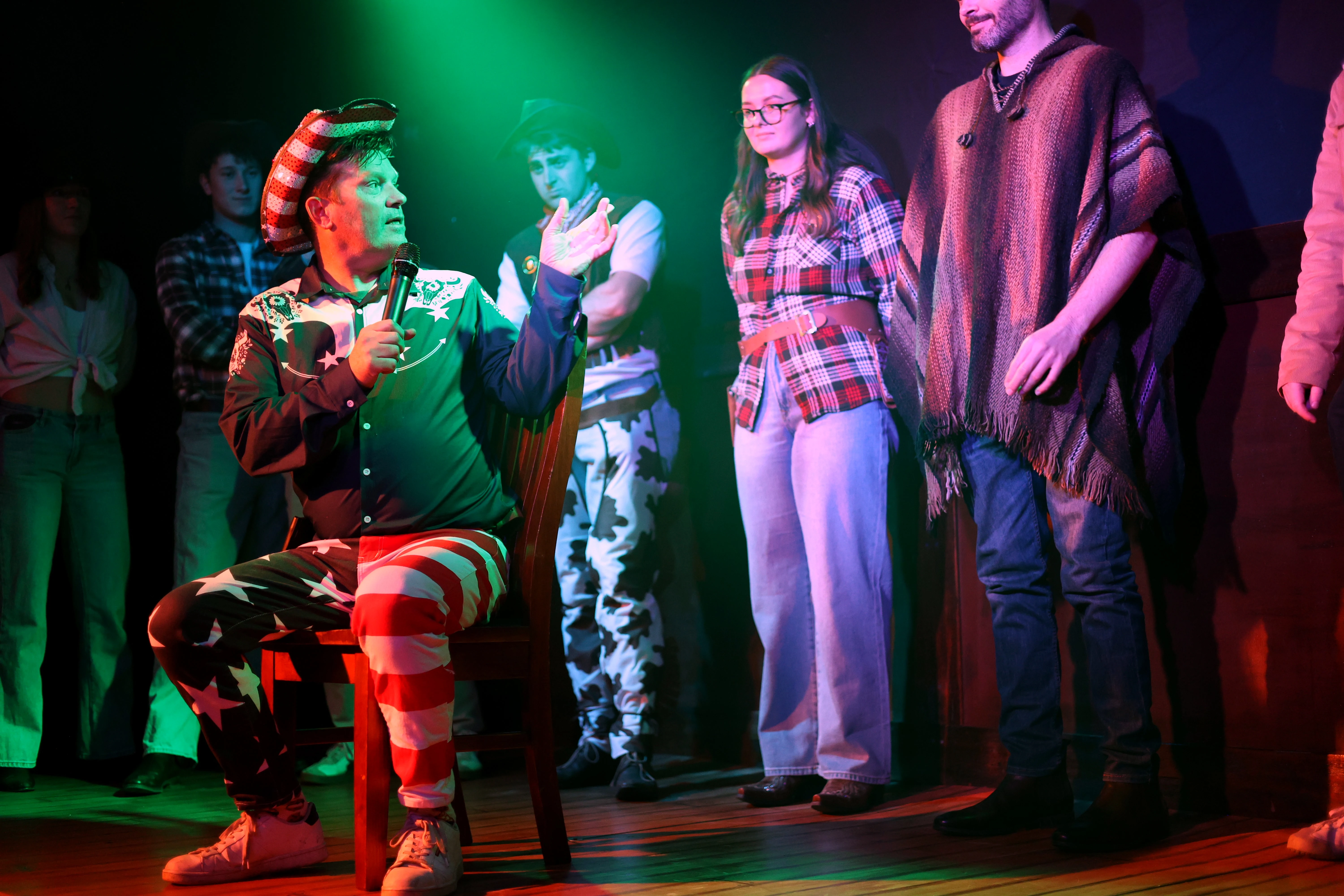 A man in a striped cowboy hat speaks into a microphone while costumed guests stand behind him