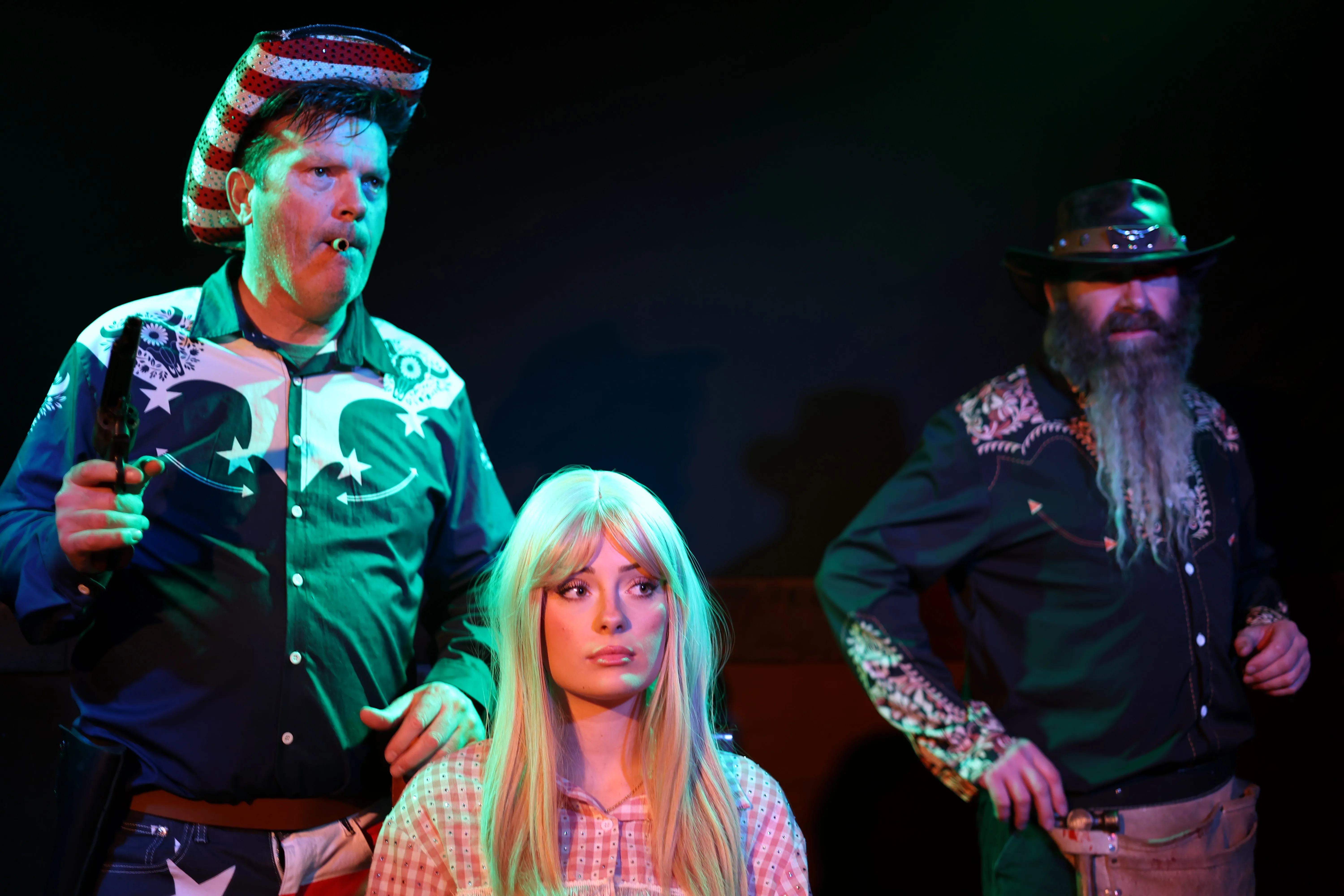 Three costumed people pose under green stage lighting, including a seated woman in a blonde wig