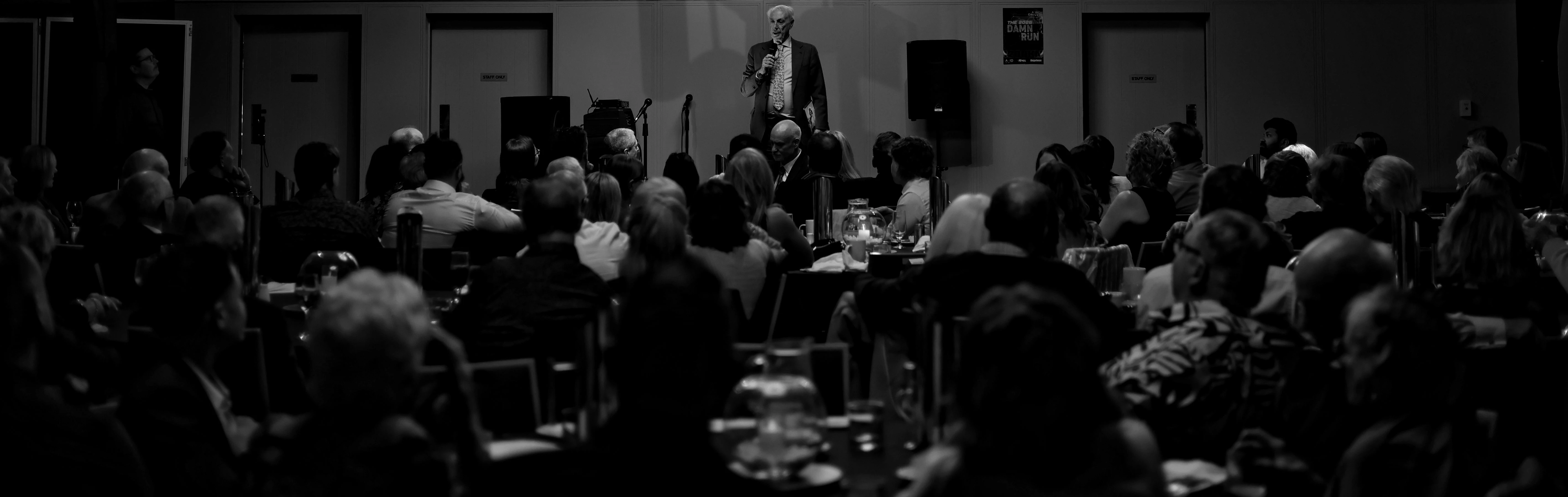 A man speaks to a crowded room in a wide black-and-white photograph