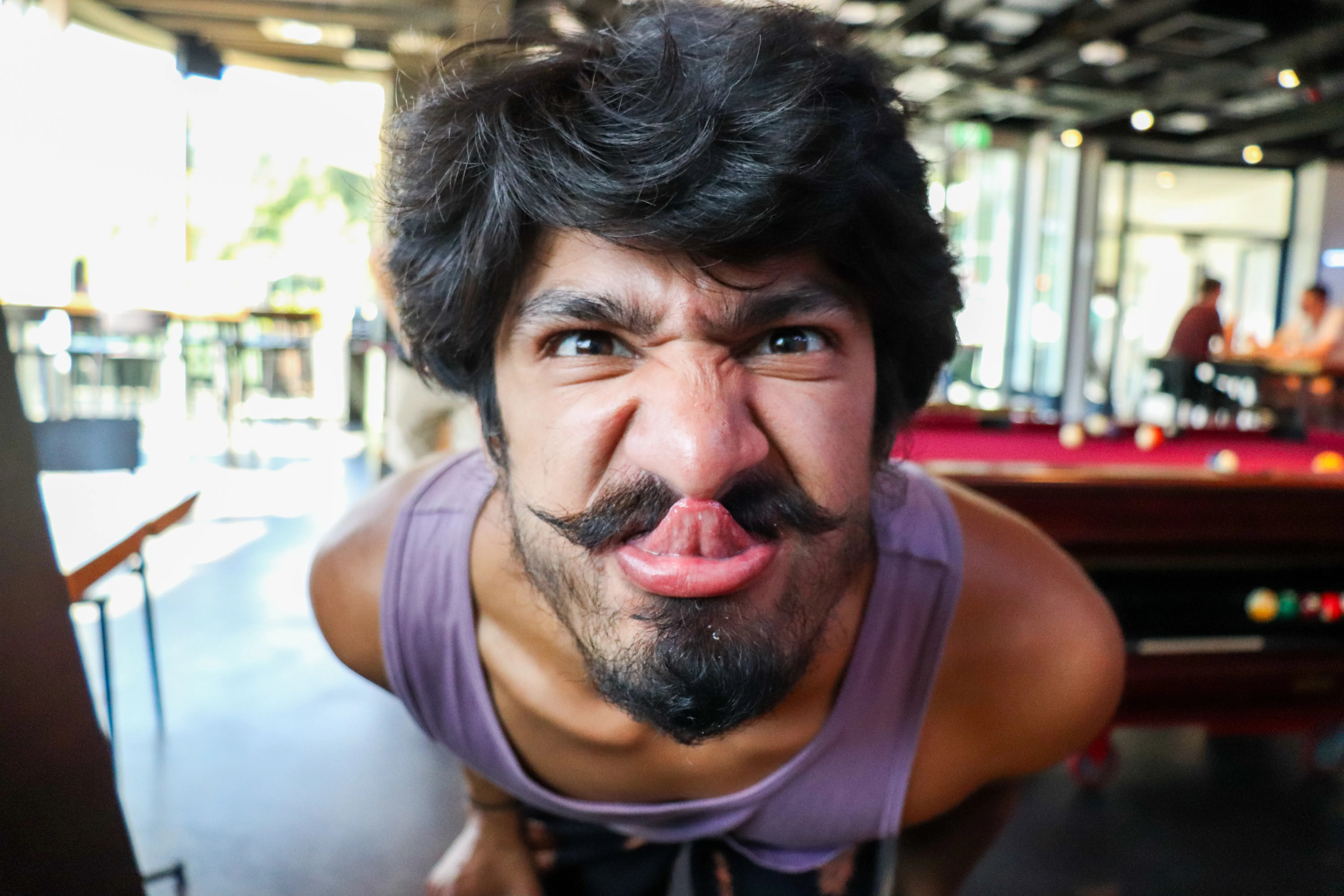 A man leans towards the camera making a playful face beside a pool table